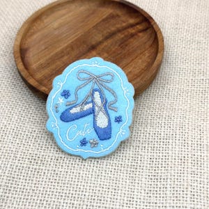 May include: A light blue embroidered patch featuring ballet shoes with silver ribbons and the word "Cute". The patch is on a wooden plate, set against a neutral fabric background.