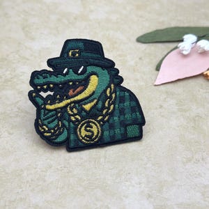 Op de afbeelding: Geborduurde patch met een groene alligator met een zwarte hoed en zonnebril, een gouden ketting en een geruit jasje. De alligator geeft een duim omhoog. De patch heeft een zwarte rand en gele accenten.