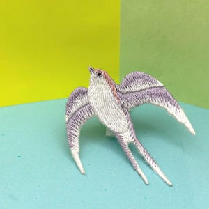 Può includere: Una rondine bianca ricamata con ali viola, posata su una superficie blu chiaro.