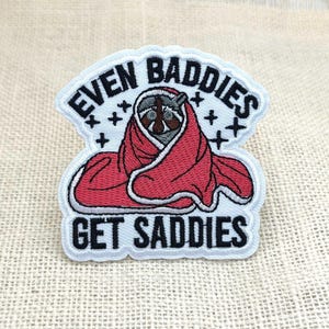 Könnte beinhalten: Gestickter Aufnäher mit einem Waschbären, der in eine rote Decke gehüllt ist. Der Aufnäher trägt den Text "EVEN BADDIES GET SADDIES" in schwarzer Schrift auf weißem Grund mit schwarzen Sternen.
