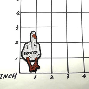 May include: White embroidered patch of a middle finger with the text "Duck You" in black. The finger is wearing a red and orange nail polish and has red and orange feet.