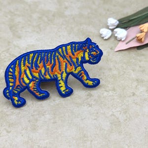 May include: Embroidered patch in the shape of a tiger. The tiger has a blue outline with yellow and orange flame-like stripes. The patch is on a light beige surface.