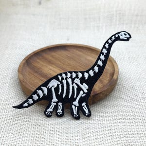 May include: A black dinosaur skeleton patch with white bone details. The patch is in the shape of a Brontosaurus, with a long neck and tail. The patch is on a wooden plate.