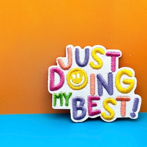 Może przedstawiać: Biała naszywka haftowana z tekstem "Just Doing My Best!" w kolorowych literach. W tekście znajduje się żółta uśmiechnięta buźka.