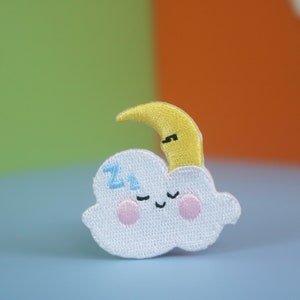 May include: A white cloud with a smiling face and the letters "zz" embroidered on it. A yellow crescent moon is on top of the cloud.