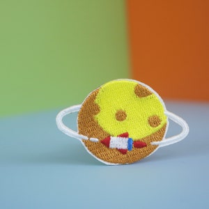 May include: A yellow embroidered planet with a white ring and a red and white rocket ship. The planet has brown spots and is surrounded by a white ring.