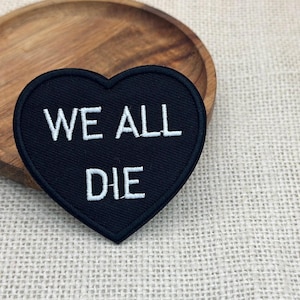 Puede incluir: Un parche negro en forma de corazón con texto bordado en blanco que dice "WE ALL DIE". El parche está colocado sobre un plato de madera, con una tela de color neutro en el fondo. El parche es una declaración simple pero audaz.