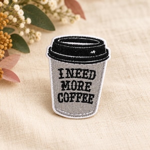 Pode incluir: Um emblema bordado em forma de copo de café descartável com tampa preta e corpo cinzento. O texto I NEED MORE COFFEE está bordado com fio preto no centro do copo. O emblema apresenta um contorno branco em toda a sua volta.