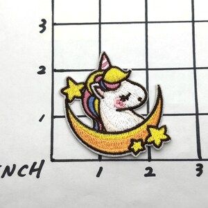 May include: A white unicorn with a yellow mane and tail is sitting on a yellow crescent moon with stars. The unicorn has closed eyes and a pink nose.