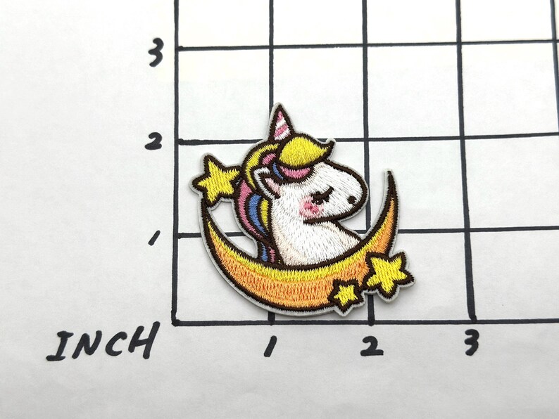 May include: A white unicorn with a yellow mane and tail is sitting on a yellow crescent moon with stars. The unicorn has closed eyes and a pink nose.