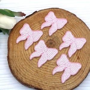 Może przedstawiać: Pięć różowych, haftowanych aplikacji w kształcie kokardek z białymi przeszyciami i różową obwódką ułożonych na drewnianym plastrze. Kokardy mają delikatny, koronkowy wygląd. Biały kwiat i zielone liście znajdują się w lewym górnym rogu.