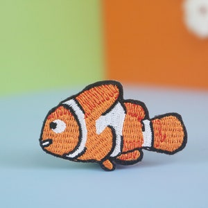 Könnte beinhalten: Ein gestickter Clownfisch-Patch in Orange und Weiß. Der Patch ist aus Stoff und hat einen schwarzen Umriss.