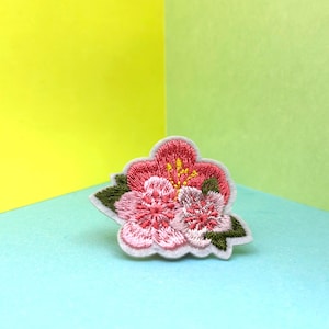 Puede incluir: Un parche bordado de flores rosas y blancas con hojas verdes. El parche está sobre un fondo azul claro.