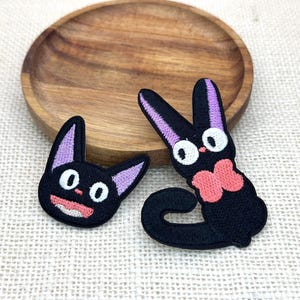 May include: Two embroidered patches featuring black cat characters. One patch shows a cat head with lavender inner ears and a pink mouth. The second patch depicts a full-body cat with lavender ears, large white eyes, and a pink bow tie at the neck.