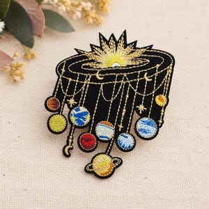 May include: A black fabric patch featuring an embroidered celestial design. The top displays a golden sunburst, while golden chains drape downward, suspending various colorful embroidered planets and small stars against the dark background.