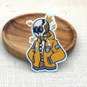 May include: An embroidered patch featuring a cartoon skull character wearing a bright yellow hooded jacket with grey accents. The character has a small orange crown above its head and is surrounded by four small orange lightning bolt symbols.