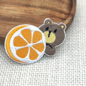 May include: Embroidered patch featuring a brown bear and a slice of orange. The bear has a yellow face and is hugging the orange slice. The orange slice is white and orange with a white border. The patch is on a textured surface.
