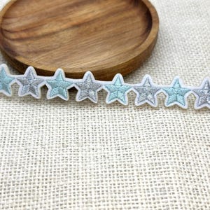 May include: A row of embroidered star patches in alternating shades of light blue and silver, outlined in white. The patches are arranged in a horizontal line. The background is a textured, off-white fabric.