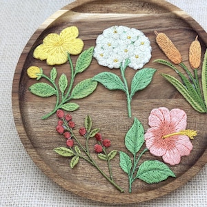 Puede incluir: Parches florales bordados en amarillo, blanco, rosa y naranja, con hojas y tallos verdes, dispuestos sobre una bandeja de madera. Los parches incluyen una flor amarilla, una hortensia blanca, un hibisco rosa y espigas.