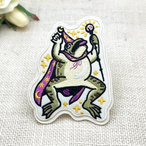May include: An embroidered patch featuring a green frog dressed as a wizard, wearing a purple cape and a pointed hat with yellow stars. The frog holds a staff topped with a glowing orb, surrounded by small yellow star accents on a white background.