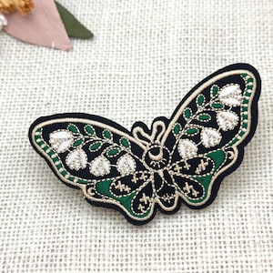 May include: Embroidered butterfly patch. The patch features a black background, green and white floral details, and gold trim. It is likely designed for clothing or accessories.