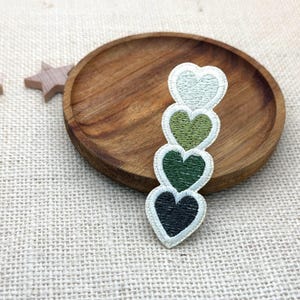May include: An embroidered patch featuring a vertical stack of four heart shapes in a gradient of green shades, ranging from a pale mint green at the top to a deep forest green at the bottom, all outlined with a light cream-colored border.