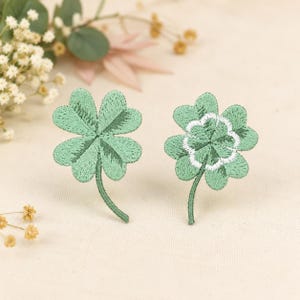May include: Two embroidered four-leaf clover patches in shades of light green. The left patch features dark green stitching details on the leaves, while the right patch includes a white circular embroidered accent in the center of the clover.