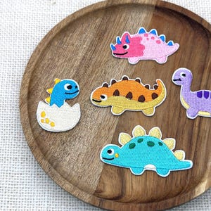 Puede incluir: Cinco parches bordados de dinosaurios en varios colores y formas. El conjunto incluye un dinosaurio azul saliendo de un huevo blanco, un triceratops rosa, un dinosaurio naranja con manchas marrones, un dinosaurio de cuello largo morado y un estegosaurio azul verdoso.