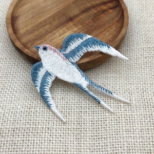 May include: Embroidered swallow patch with blue and white wings, a white body, and a pink head. The bird patch is on a textured, off-white surface, with a wooden bowl in the background.