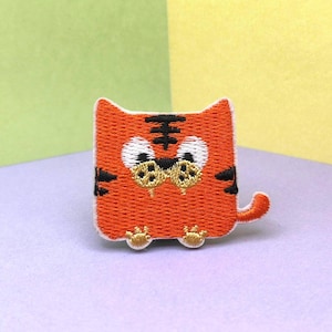 以下が含まれることがあります： 黒い縞模様と白い目を持つ、刺繍されたオレンジ色のトラのパッチ。パッチは小さな尾と小さな足を持つ正方形の形をしています。この装飾的なパッチは、衣類やアクセサリーにアイロンをかけたり、縫い付けたりすることができます。