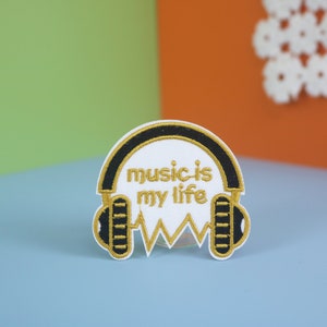 Puede incluir: Parche bordado con diseño de auriculares blancos con detalles negros y dorados. El texto "music is my life" está centrado. Incluye un diseño de onda de sonido dorada en la parte inferior.
