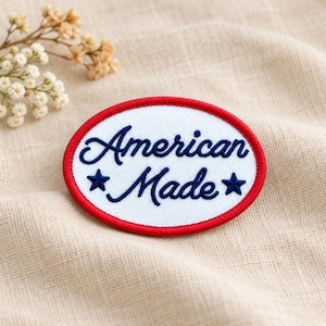 May include: An oval-shaped embroidered patch with a white background and a red stitched border. The text American Made is embroidered in dark blue cursive script, flanked by two small dark blue stars on either side of the word Made.