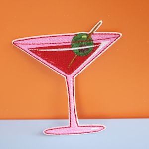 Peut inclure: Écusson brodé représentant un verre à martini rose rempli d'une boisson rouge, orné d'une olive verte sur un cure-dent. Le patch a une bordure blanche et est posé sur un fond orange. Le design est détaillé et coloré.