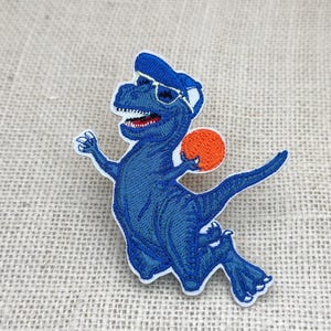 May include: Embroidered patch featuring a blue dinosaur wearing a cap and sunglasses, holding an orange ball. The dinosaur has white teeth and is outlined in white. The patch is on a textured, neutral-colored background.