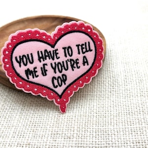 May include: A heart-shaped patch with the text "YOU HAVE TO TELL ME IF YOU'RE A COP" in black. The patch has a pink center, a red border, and a scalloped edge. It rests on a wooden dish.