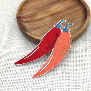 Puede incluir: Dos parches bordados de pimientos. Uno es rojo, el otro naranja, ambos con contornos blancos y tallos azules. Los parches están sobre una superficie blanca texturizada, cerca de un plato de madera.