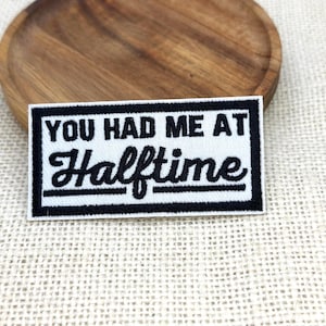 May include: A rectangular patch with the text "YOU HAD ME AT Halftime" in black and white. The patch has a black border and is on a textured, off-white background. The patch is placed on a wooden plate.