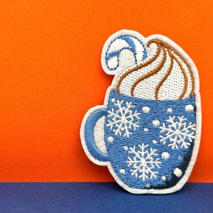 Pode incluir: Um remendo bordado azul e branco de uma caneca com chantilly e um bastão de caramelo no topo. A caneca é decorada com flocos de neve e pontos brancos.