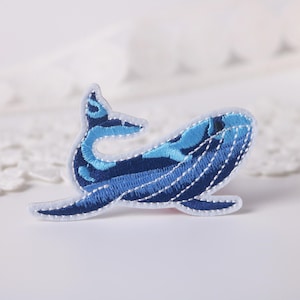May include: Embroidered whale patch in shades of blue and white. The whale design features intricate stitching details, with a dark blue body and lighter blue accents. The patch is on a white background.