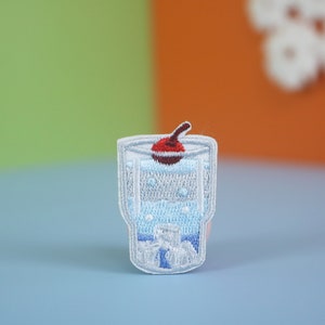 Könnte beinhalten: Ein weißer gestickter Aufnäher mit einem Glas mit einer Kirsche oben drauf. Das Glas ist mit einer blauen Flüssigkeit gefüllt und hat Blasen.