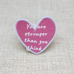 Peut inclure: Un écusson en forme de cœur rose avec le texte brodé blanc « You are stronger than you think ». L'écusson a une bordure blanche et est posé sur un fond texturé de couleur neutre.