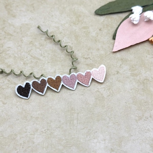 May include: A series of heart-shaped embroidered patches in shades of brown and pink, each with a white border. The patches are arranged in a slight curve. Decorative floral elements are in the background.