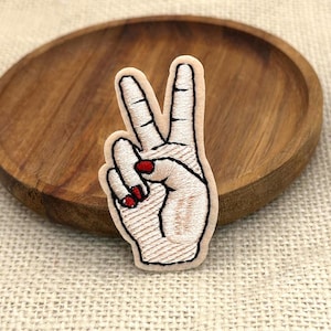 May include: An embroidered patch featuring a hand making a peace sign gesture. The hand is stitched in light beige thread with black outlines and details. The fingernails are filled with bright red thread, creating a distinct contrast.