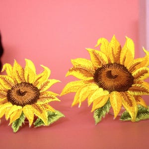 Könnte beinhalten: Zwei gestickte Sonnenblumen-Patches, einer größer als der andere, mit gelben Blütenblättern und braunen Zentren. Die Patches befinden sich auf einem rosa Hintergrund.