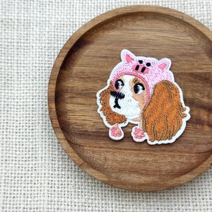 May include: Embroidered patch featuring a dog wearing a pink pig hat. The dog has brown and white fur with long, floppy ears. The patch is on a wooden plate with a light-colored fabric background.