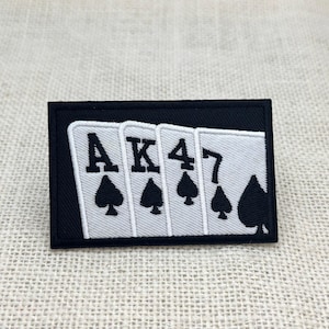 May include: A rectangular black patch featuring four playing cards: Ace, King, Four, and Seven of Spades. The cards are white with black spade symbols and text. The patch is likely for clothing or accessories.