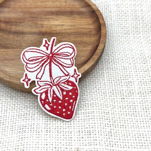 May include: Embroidered patch featuring a red strawberry with a bow and stars. The strawberry has a textured appearance with a white outline. The patch is on a white background with a wooden plate.