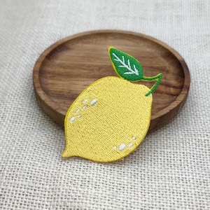 Könnte beinhalten: Gestickter gelber Zitronenaufnäher mit grünem und weißem Blatt. Der Aufnäher liegt auf einem kleinen, runden Holztablett vor einem neutralen Leinenhintergrund. Die Zitrone hat weiße Stickdetails.