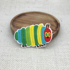 May include: An embroidered patch shaped like a caterpillar with a red head, yellow eyes, and a segmented body featuring alternating vertical stripes of green, yellow, and blue. The patch has a white border and small brown feet along the bottom edge.