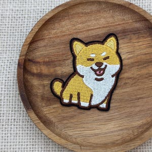 Op de afbeelding: Geborduurde patch met een lachende Shiba Inu hond. De patch heeft een geel en wit kleurenschema met zwarte contouren. De hond zit met zijn tong uit. De patch ligt op een houten bord.